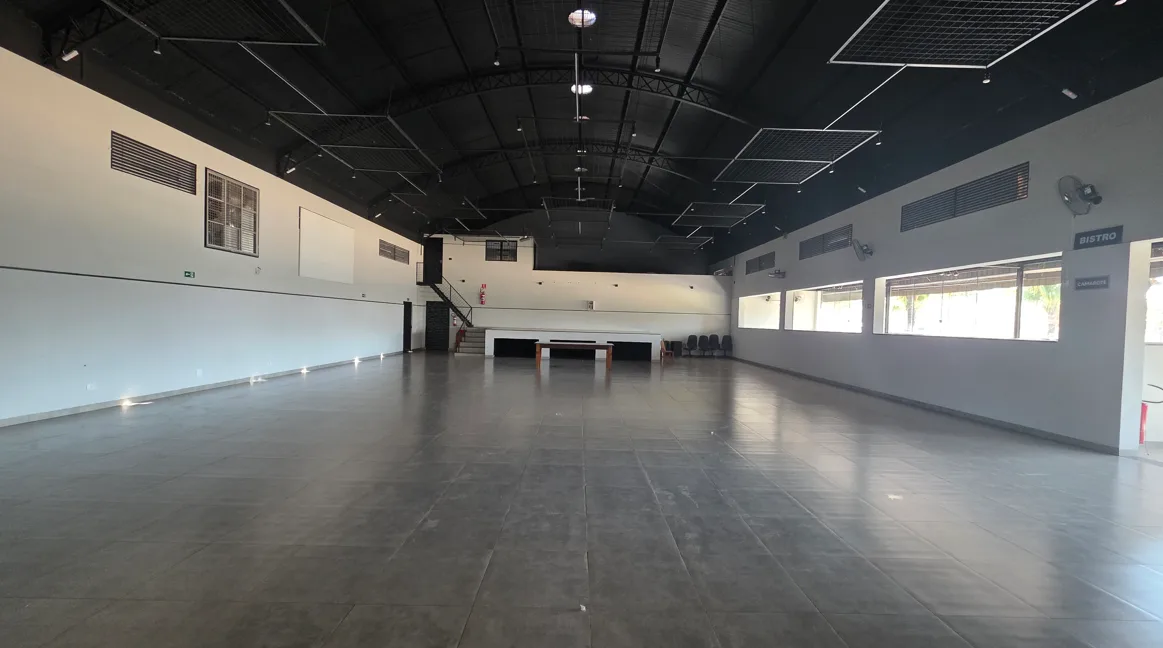 Salão amplo da AK Eventos com piso claro, teto metálico com iluminação, escada lateral, áreas de apoio e palco branco ao fundo